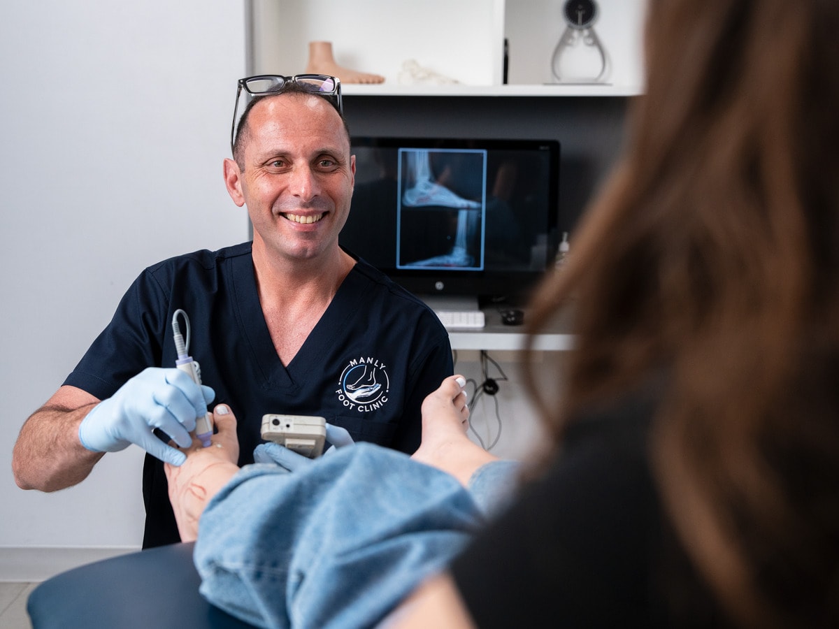 Dr Ozan Amir, Surgical Podiatrist, with a handheld Doppler at a patient’s foot and a foot X-ray on the screen behind him