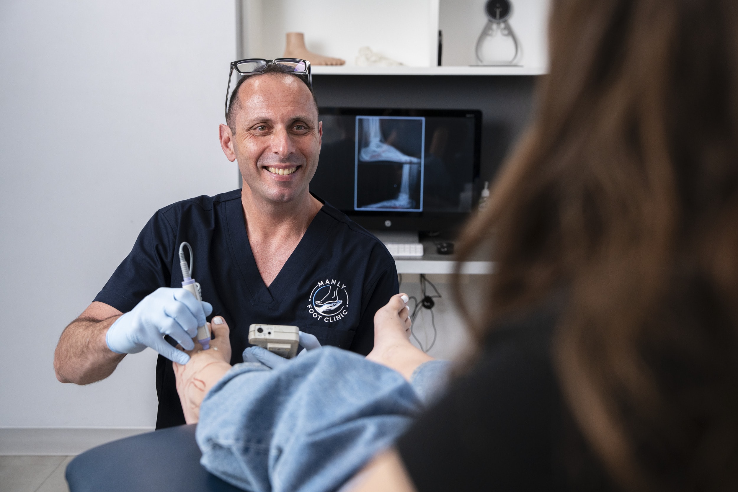 Ozan Amir, surgical podiatrist, checking the circulation in a patient’s foot with a handheld Doppler at Manly Foot Clinic, a foot X-ray on the screen behind him